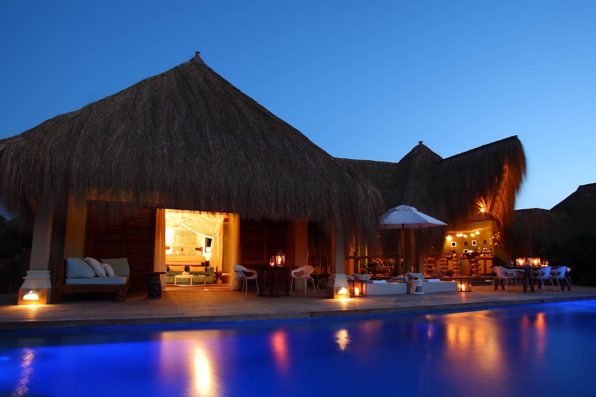 Thatched-roof bungalows with warm lighting overlook an illuminated pool at dusk at Azura Benguerra Island.