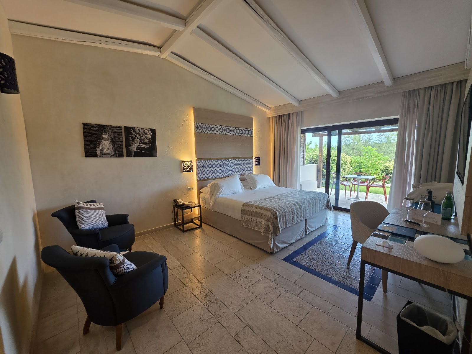 Spacious resort bedroom with king bed, black armchairs, desk area, and sliding glass doors opening to a balcony overlooking greenery.