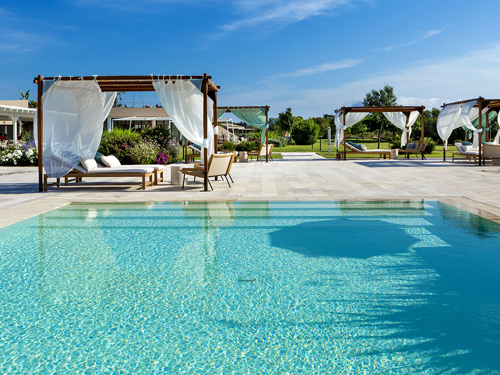 Swimming pool with clear turquoise water bordered by white stone deck, wooden pergolas with flowing white curtains, lounge chairs, and manicured grounds under blue sky.