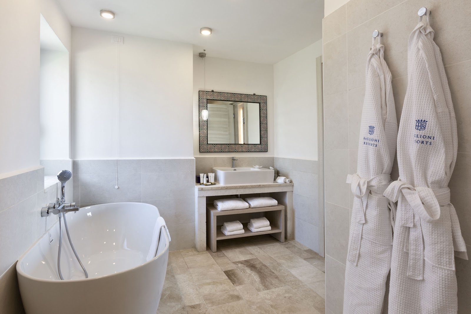 Spacious bathroom at Baglioni Resort with freestanding oval tub, vessel sink, framed mirror, and monogrammed bathrobes hanging on wall hooks.