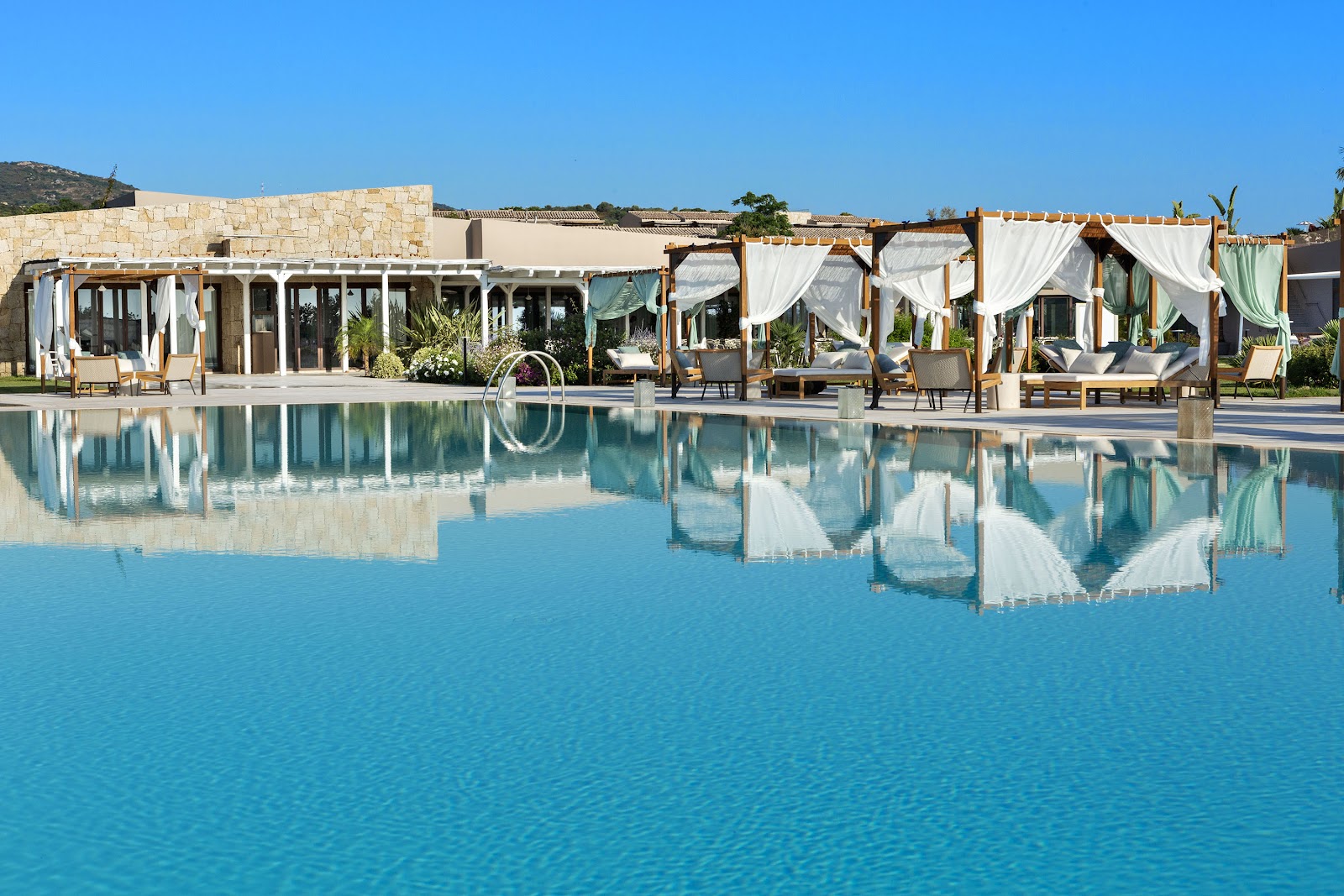 Resort pool with draped wooden cabanas, loungers, and stone buildings under clear blue sky.