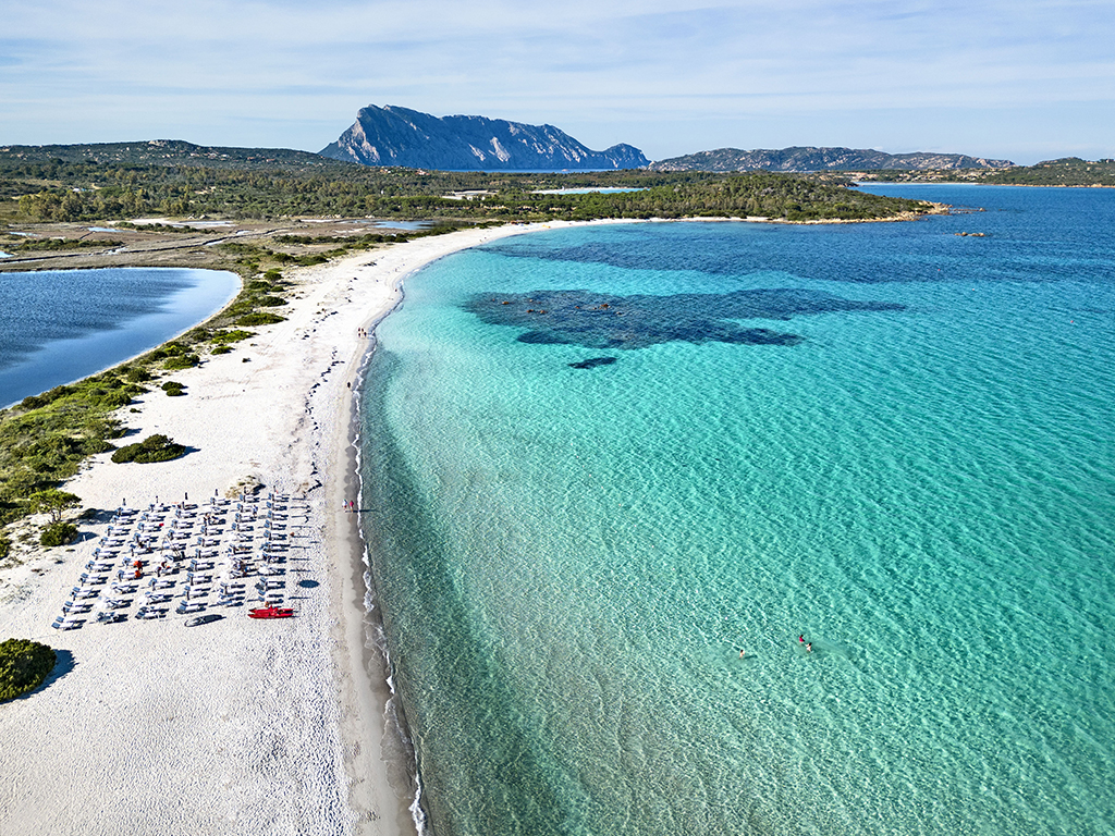 White sand beach with blue umbrellas and loungers at Baglioni Resort, turquoise waters and rocky mountain backdrop visible across the bay.