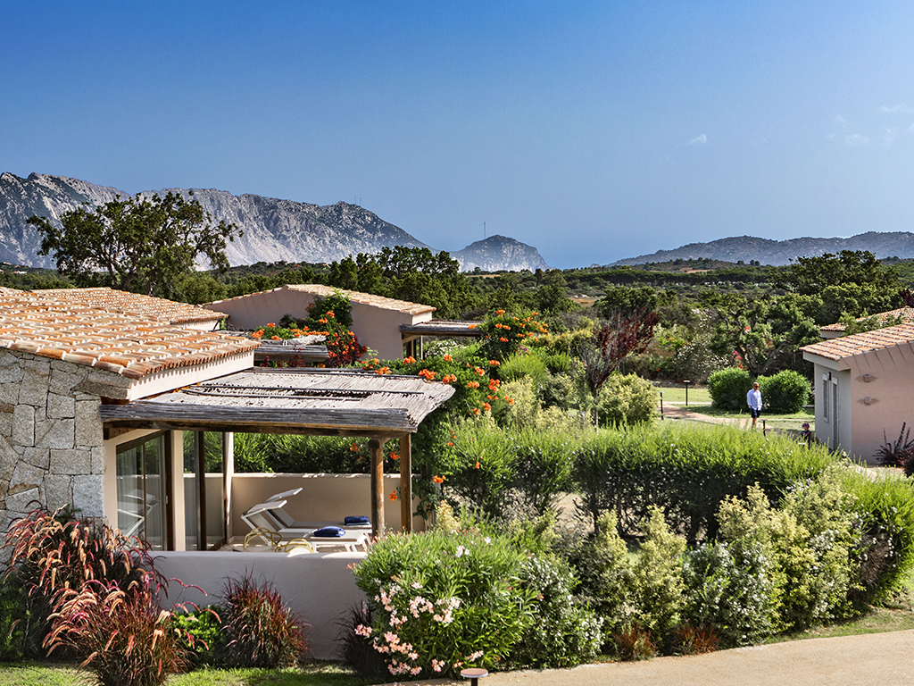 Beige bungalow with terracotta roof and covered patio surrounded by lush gardens, mountains visible across the valley under clear sky.