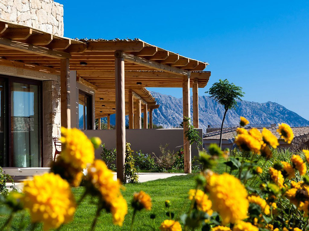 Wooden pergola with stone building overlooks manicured lawn and yellow flowers, with mountains visible in the distance under clear blue sky.