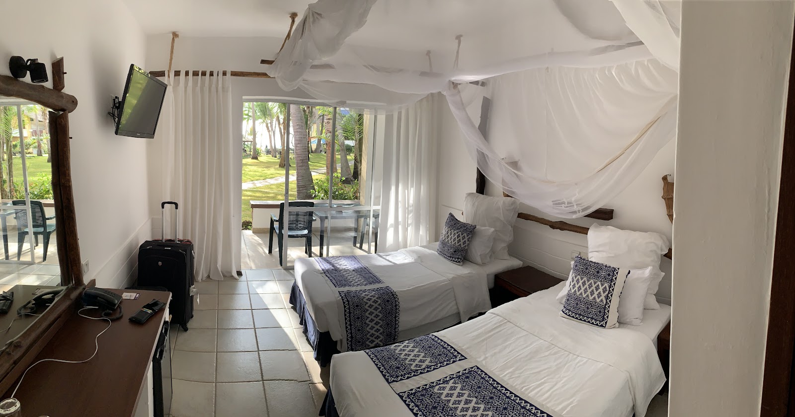 Resort room with two beds topped with white linens and blue patterned runners, sheer canopy drapes, desk, and glass doors opening onto a tropical garden with palm trees.