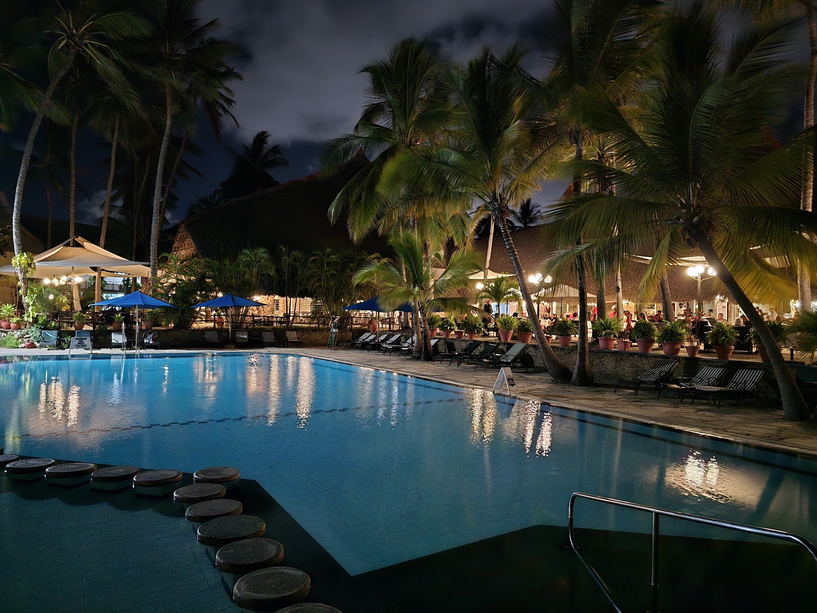 Resort pool lit at dusk with palm trees, lounge chairs, umbrellas, and lit buildings along the waterfront at Bahari Beach Hotel.