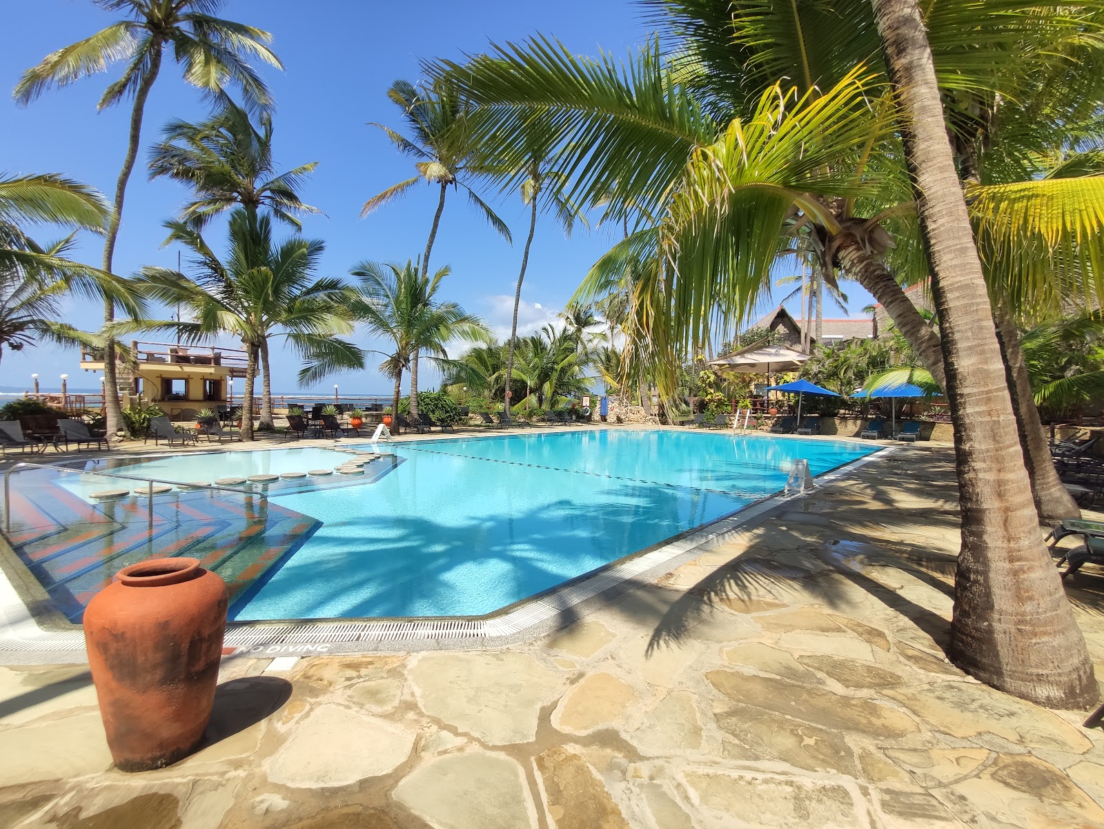 Resort pool surrounded by tall palm trees, with sandy beach, blue umbrellas, and ocean view in background at Bahari Beach Hotel.