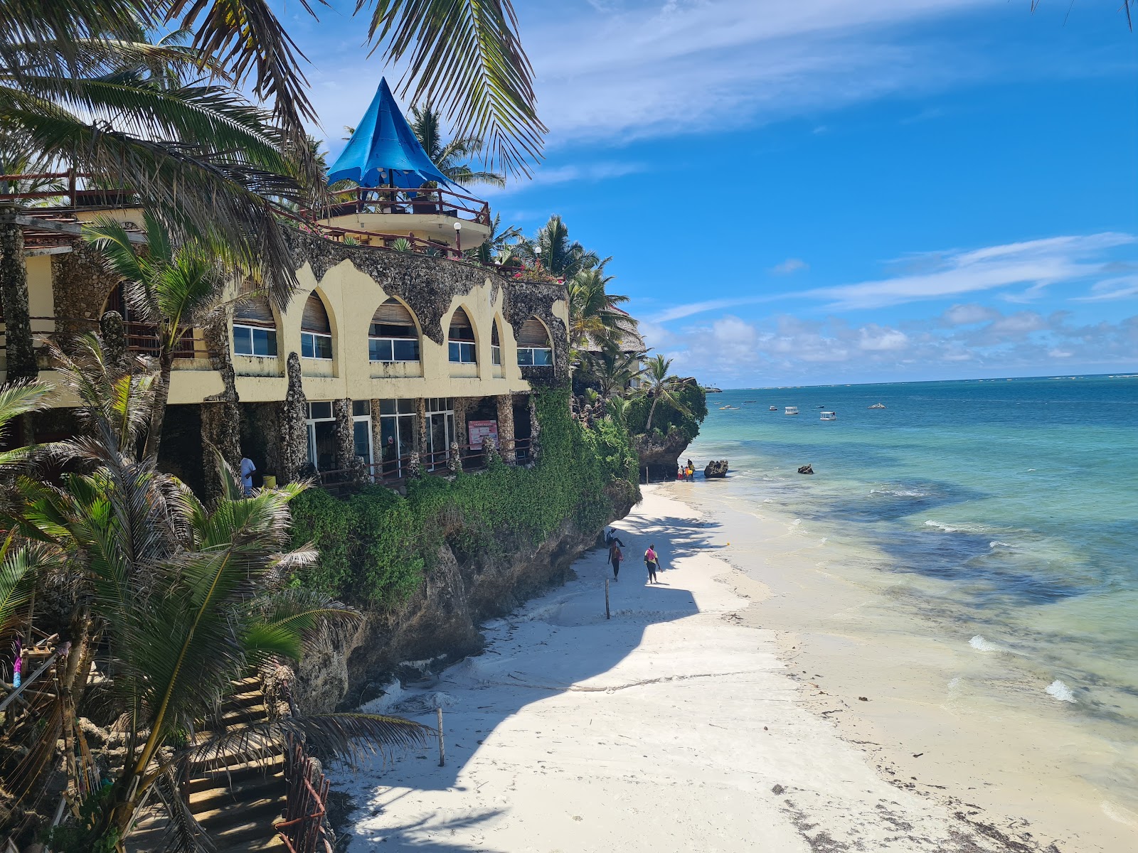 Bahari Beach Hotel's cream-colored building with arched windows sits on a rocky cliff above a white sand beach, with a blue umbrella on the rooftop deck and turquoise ocean beyond.