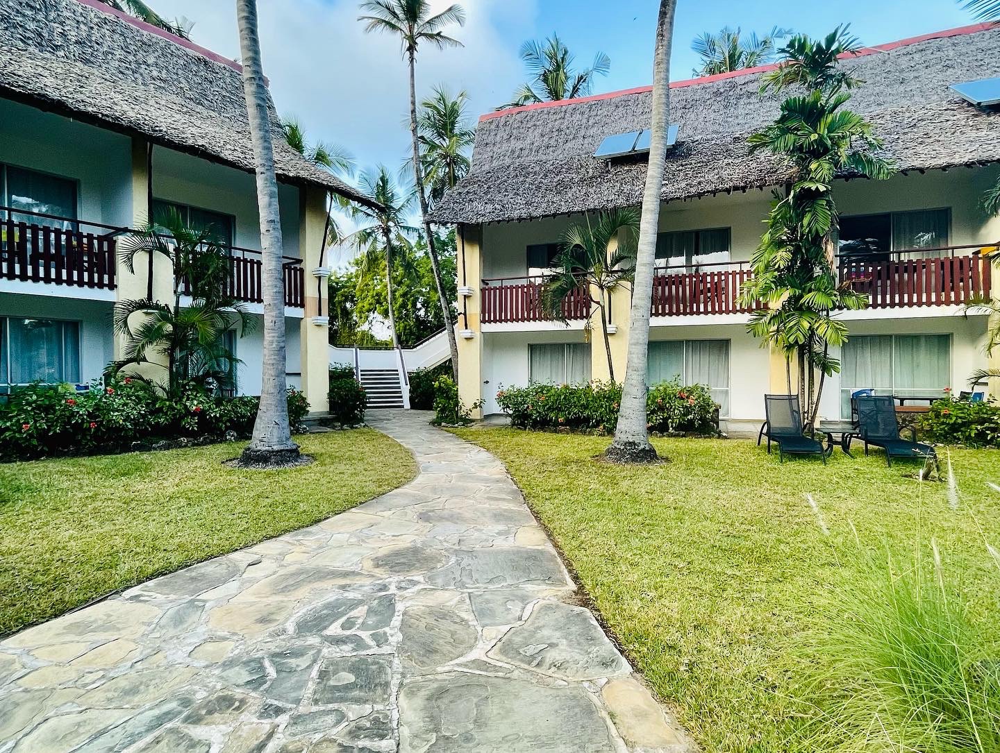 Stone pathway lined with palm trees leads between thatched-roof bungalows with wooden balconies at Bahari Beach Hotel.