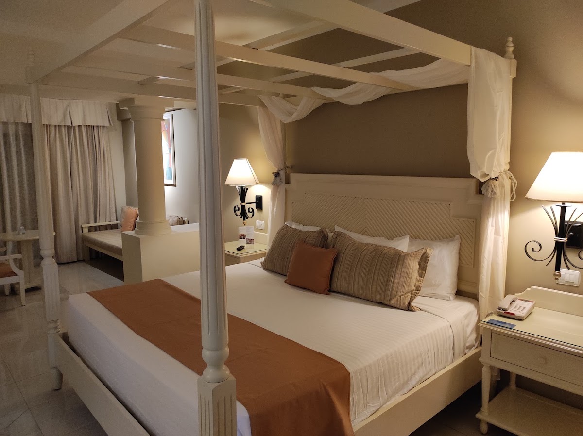 Four-poster canopy bed with cream linens and striped pillows in a neutral bedroom with sitting area and vanity.