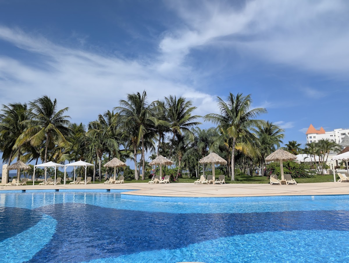 Blue resort pool with loungers and thatched umbrellas along the deck, palm trees, and white building with orange roof in background.