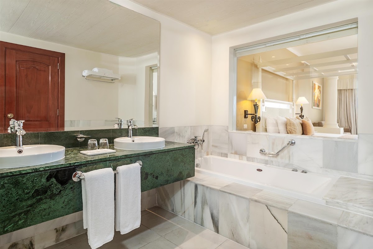 Resort bathroom with green marble vanity, dual white sinks, soaking tub, and view into bedroom through open doorway.