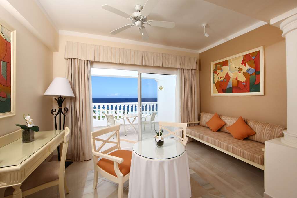 Resort suite with ocean view from balcony, striped sofa with orange pillows, round table, and coastal furnishings in warm tones.