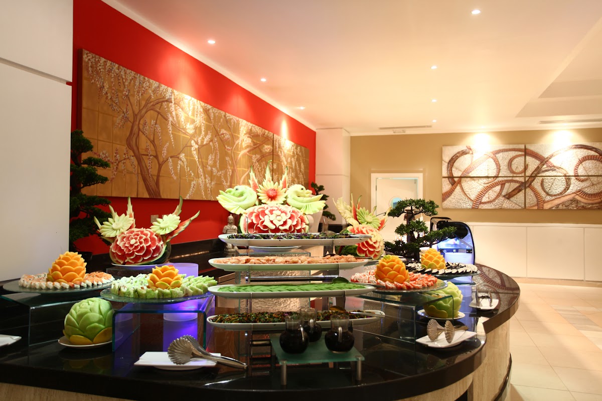 Curved black buffet table displays carved fruit sculptures and vegetable arrangements under warm lighting, with red accent wall and golden tree artwork behind.