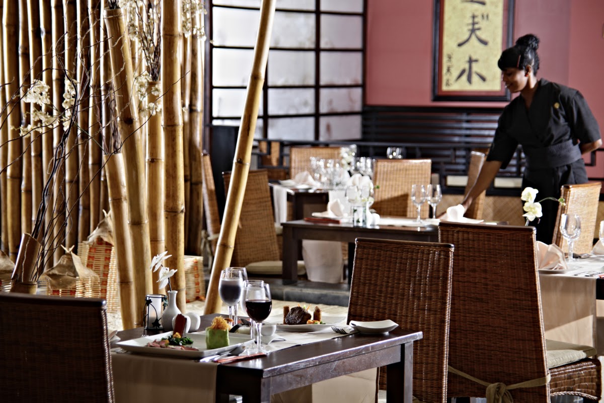 Fine dining room with wicker chairs, plated appetizers, wine glasses, and tall bamboo stalks with white flowers at Bahia Principe Escape Runaway Bay.