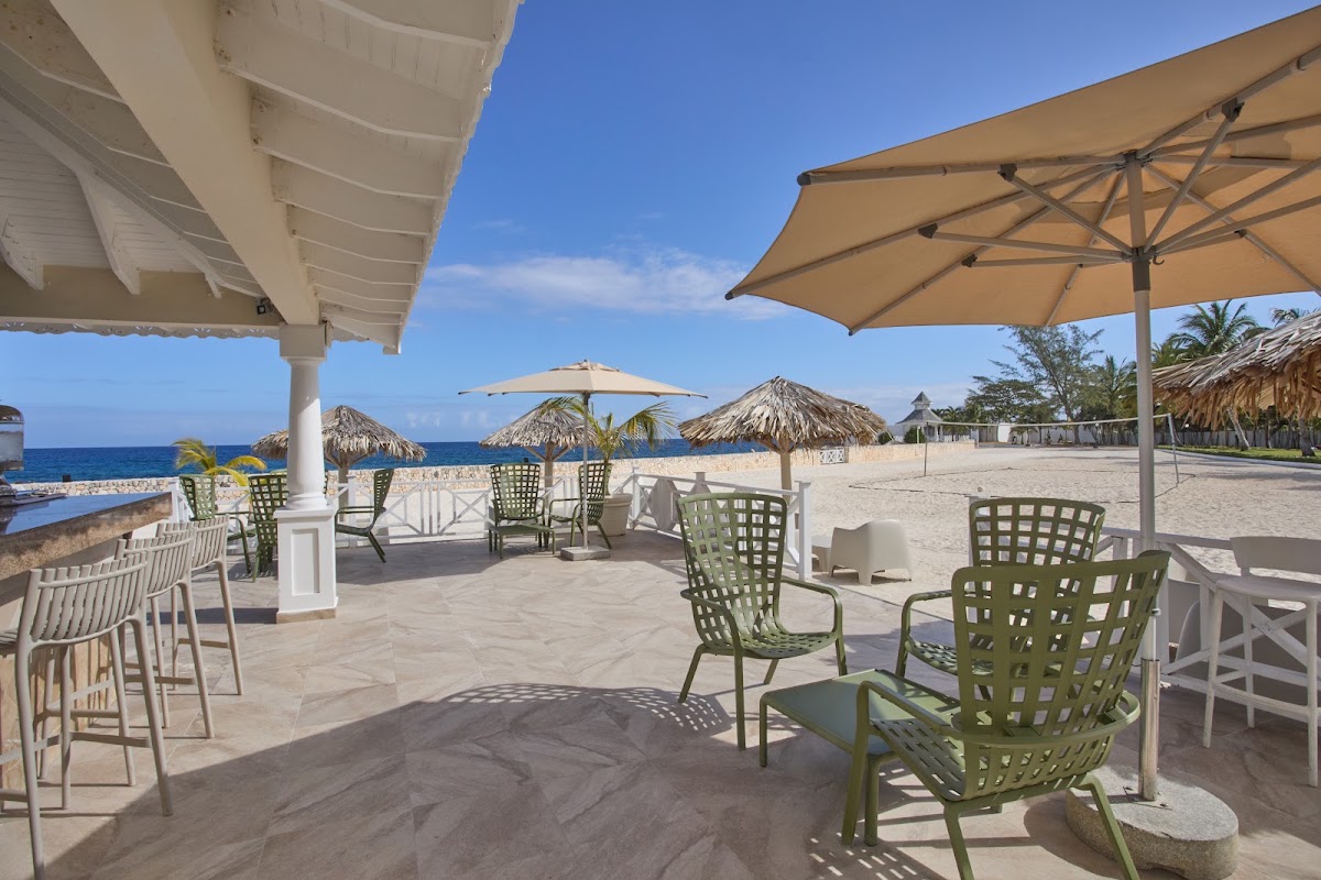 Shaded seating area with green and white chairs overlooks a sandy beach with thatched umbrellas and the ocean beyond at Bahia Principe Escape Runaway Bay.
