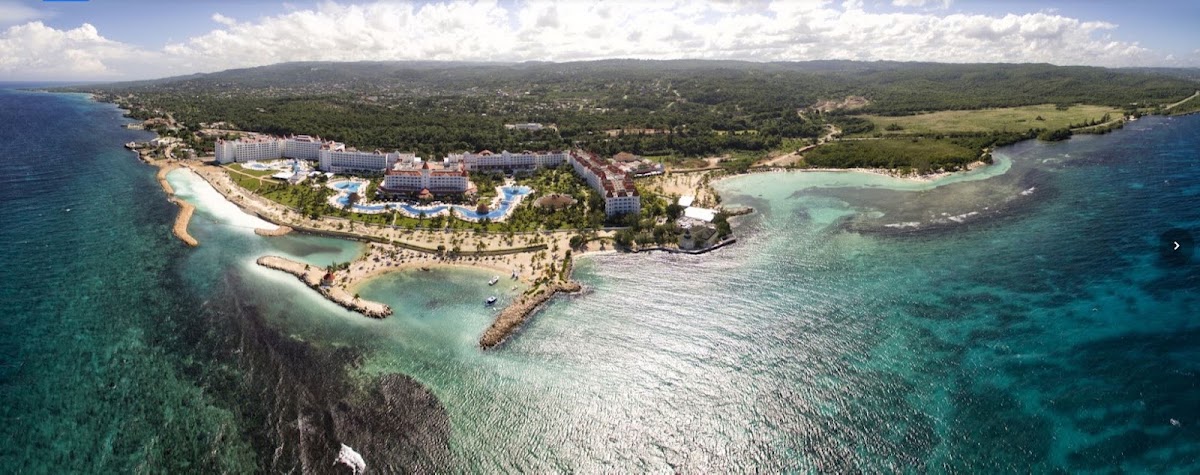Aerial view of Bahia Principe Escape Runaway Bay nestled on a curved coastline with turquoise waters, white sand beaches, and forested hills beyond.