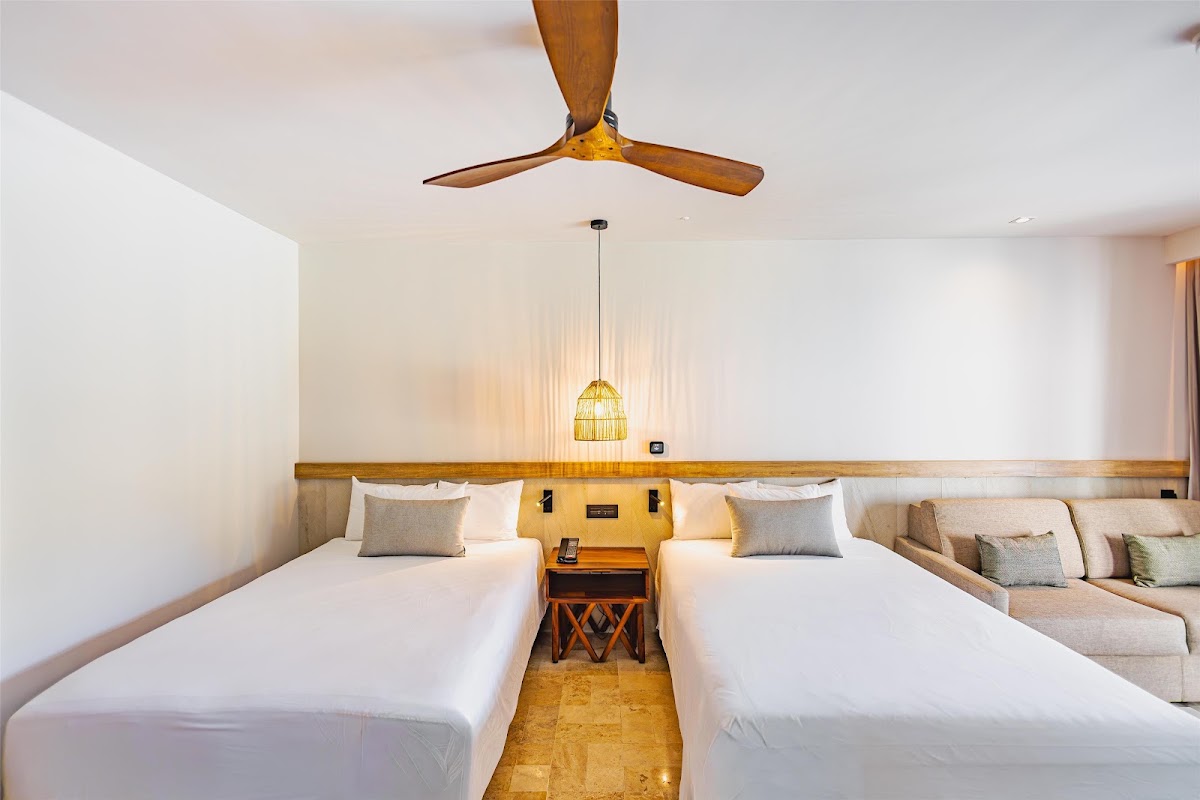 Two queen beds with white linens and cushions face each other in a modern resort room with wooden ceiling fan and pendant lighting.