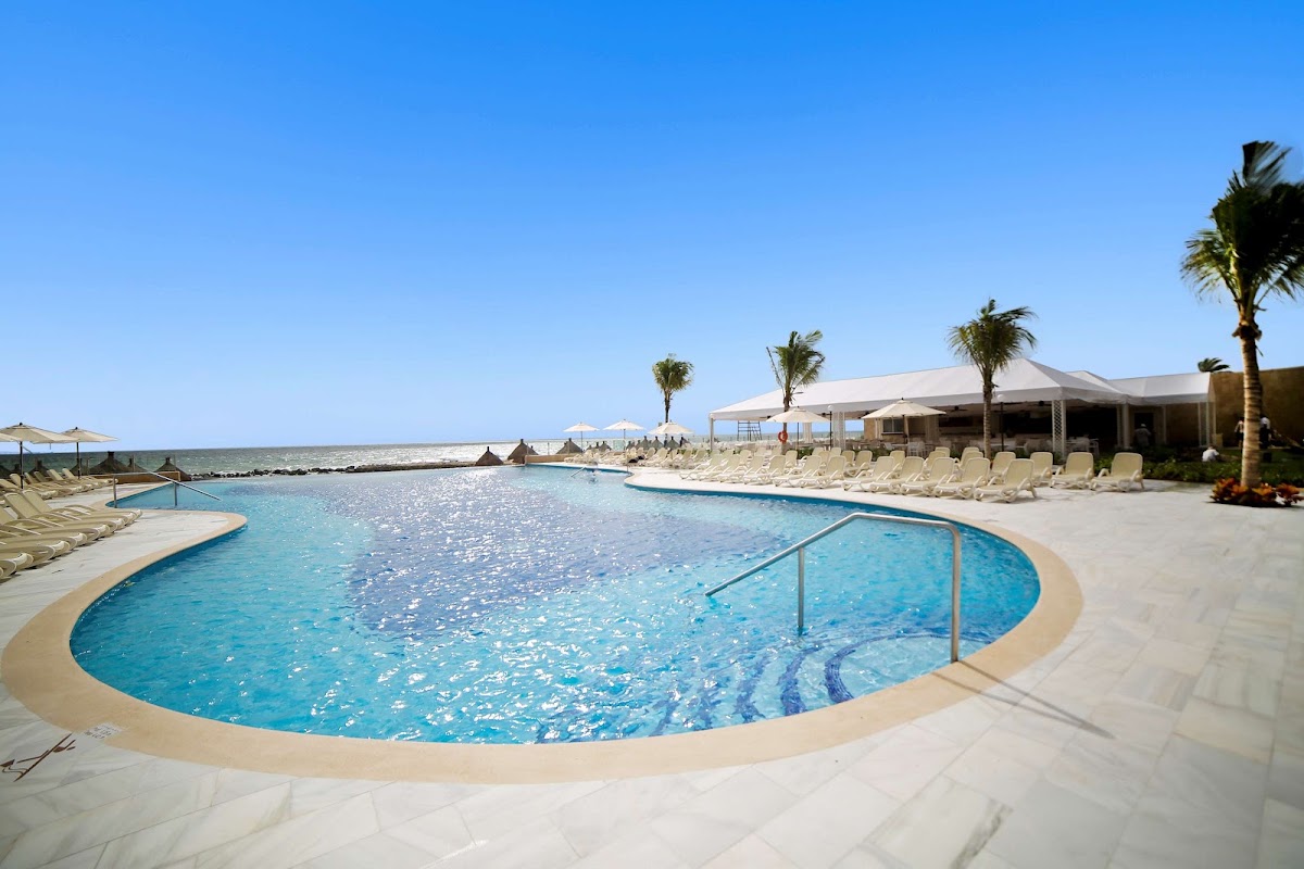 Resort pool with lounge chairs, umbrellas, and palm trees overlooking the beach and sea under clear blue sky.