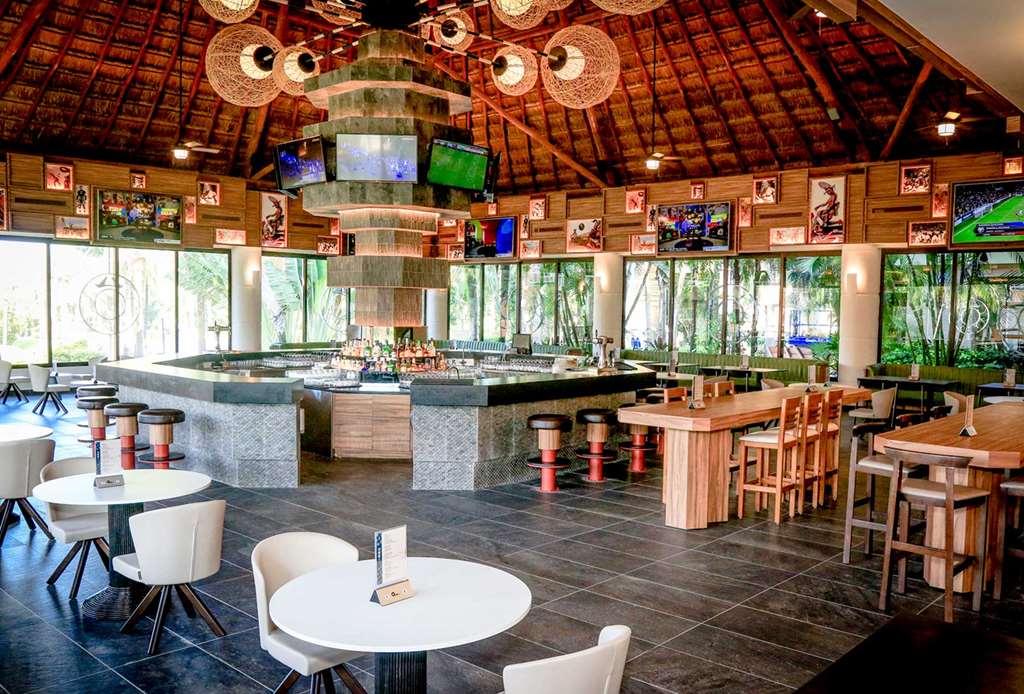 Open-air bar and lounge with thatch roof, central bar counter, wooden dining tables, white round tables, TV screens, and forest views through large windows.