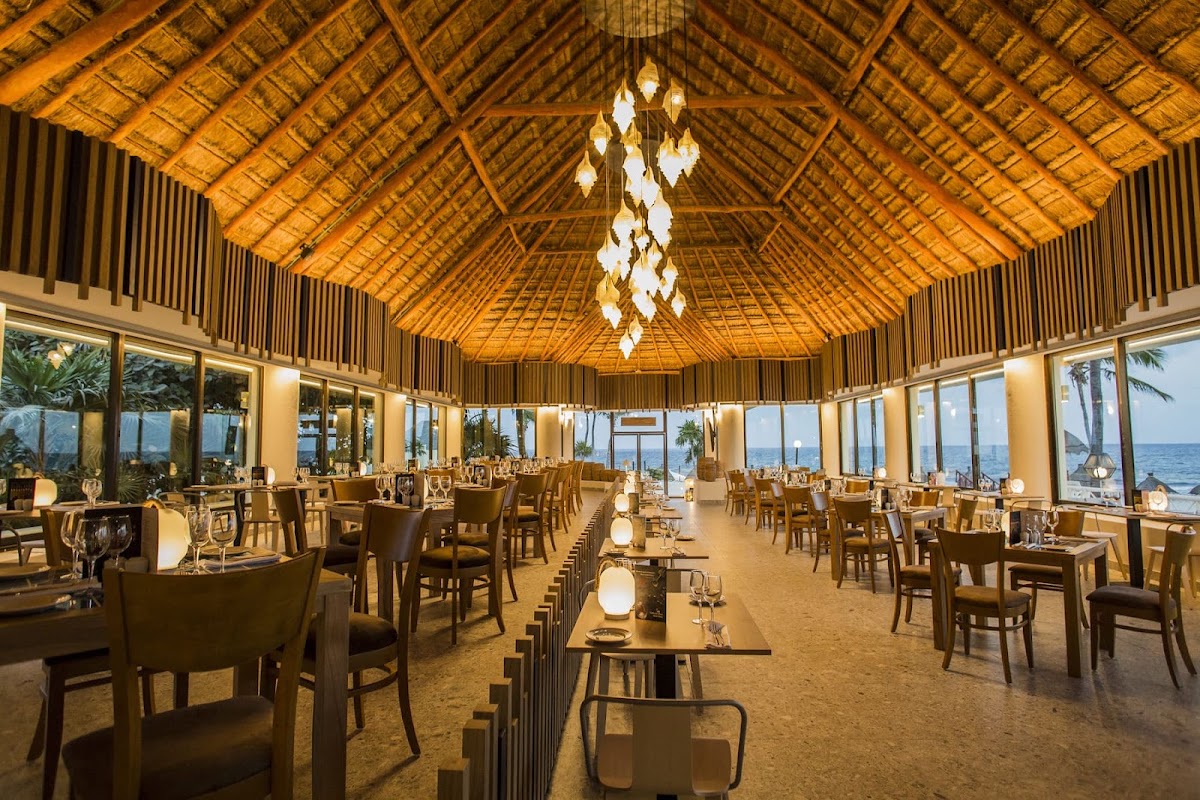 Dining room with thatched roof, wooden beams, leaf-shaped chandelier, and floor-to-ceiling windows overlooking the ocean at Bahia Principe Explore Akumal.