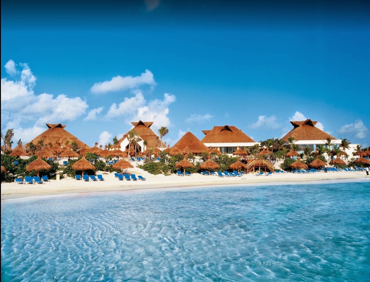 Beach view of Bahia Principe Explore Akumal with thatched-roof buildings, blue loungers, and clear turquoise water.