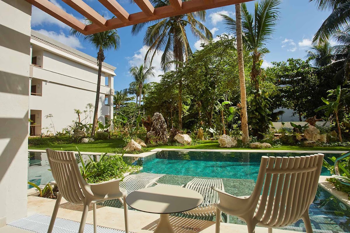 Two lounge chairs overlook a turquoise pool surrounded by manicured lawn, palm trees, and a modern white building at Bahia Principe Explore Akumal.