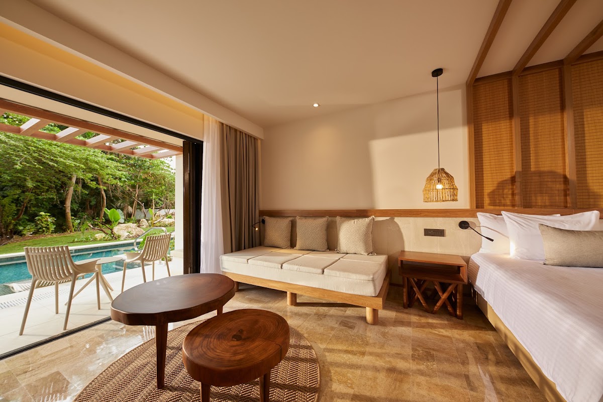 Resort suite with wooden furniture, sofa, and bed opening to a terrace with pool and tropical trees.