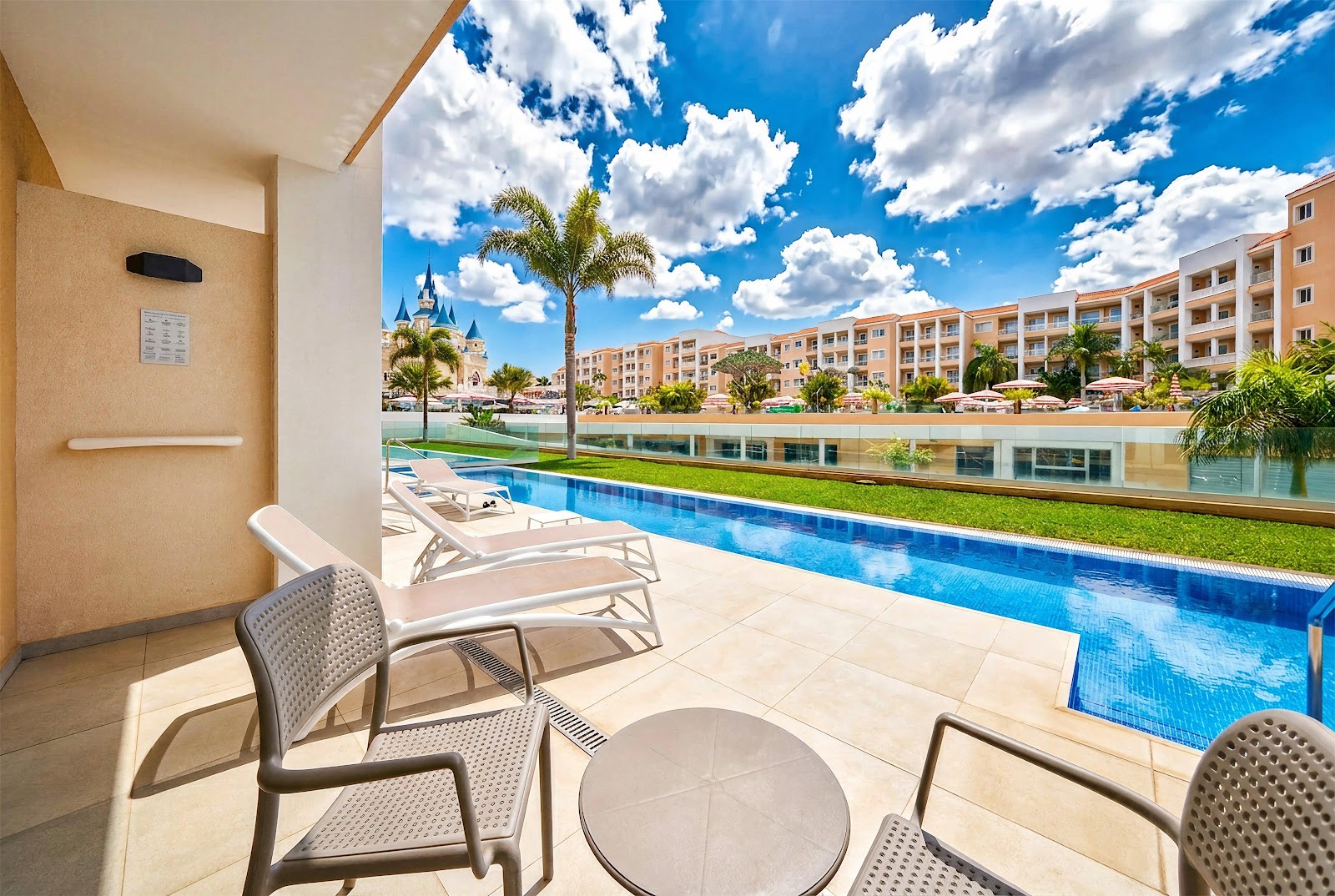 Lounge chairs and table on a patio overlooking a blue pool, manicured lawn, and multi-story resort buildings with a castle visible in the distance.
