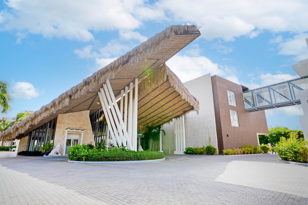 Modern resort entrance with thatched roof structure, white columns, and brick building under blue sky with clouds.