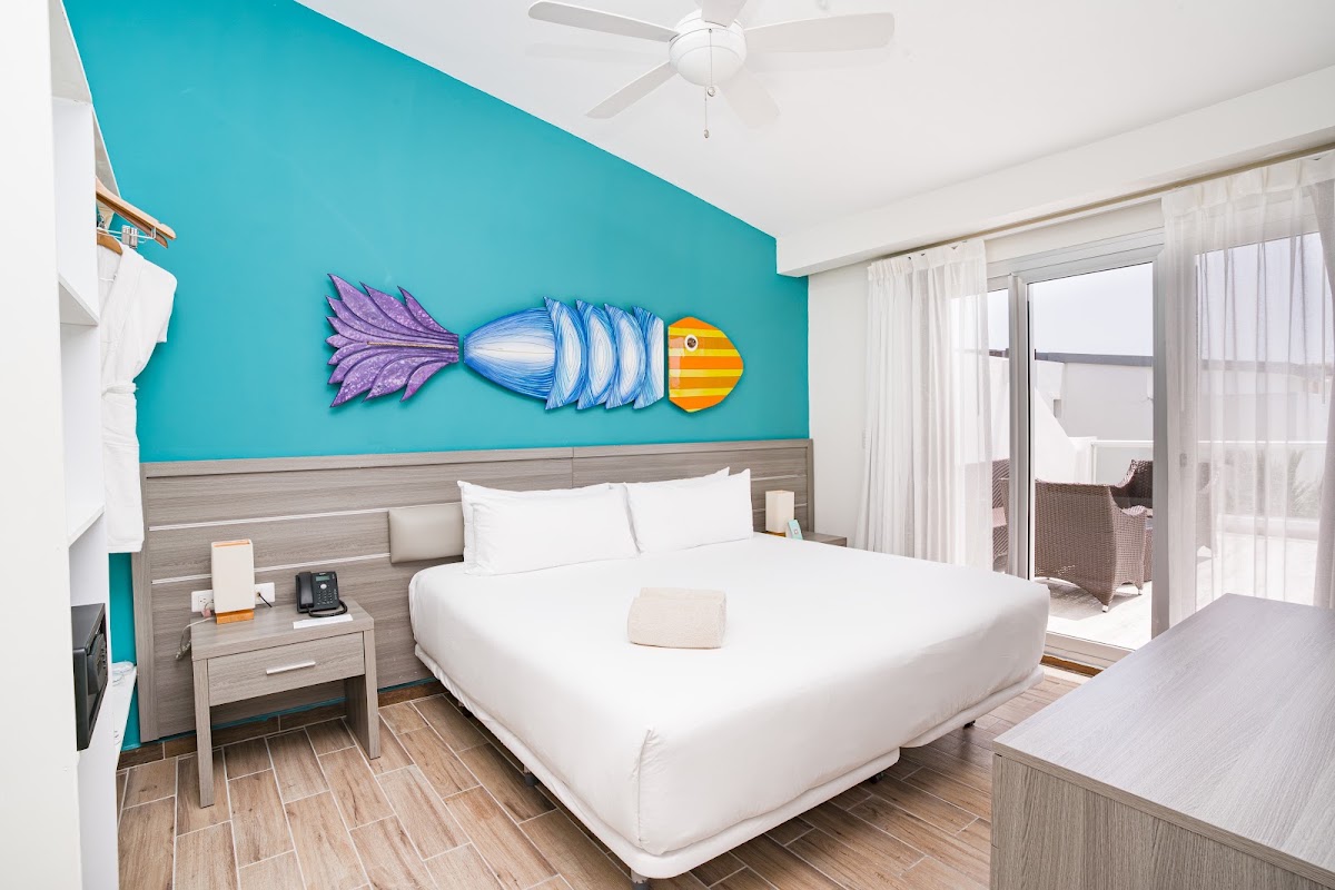 Bright resort bedroom with turquoise accent wall featuring colorful fish skeleton art above the bed, white bedding, and balcony access.