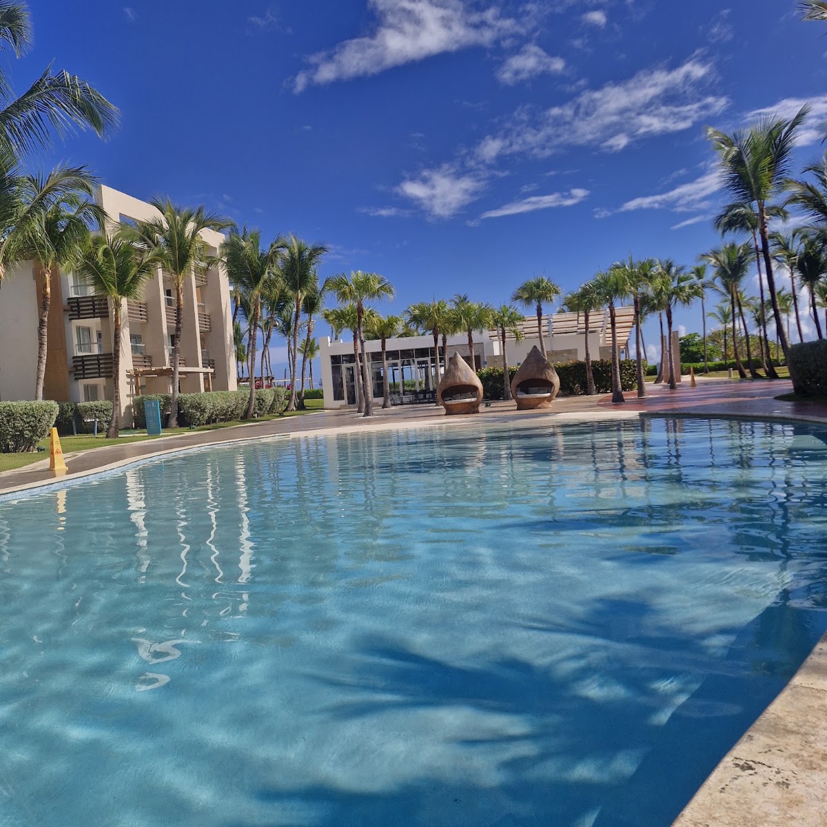 Resort pool with turquoise water, surrounded by palm trees, beige building, and thatched-roof palapas under clear blue sky.