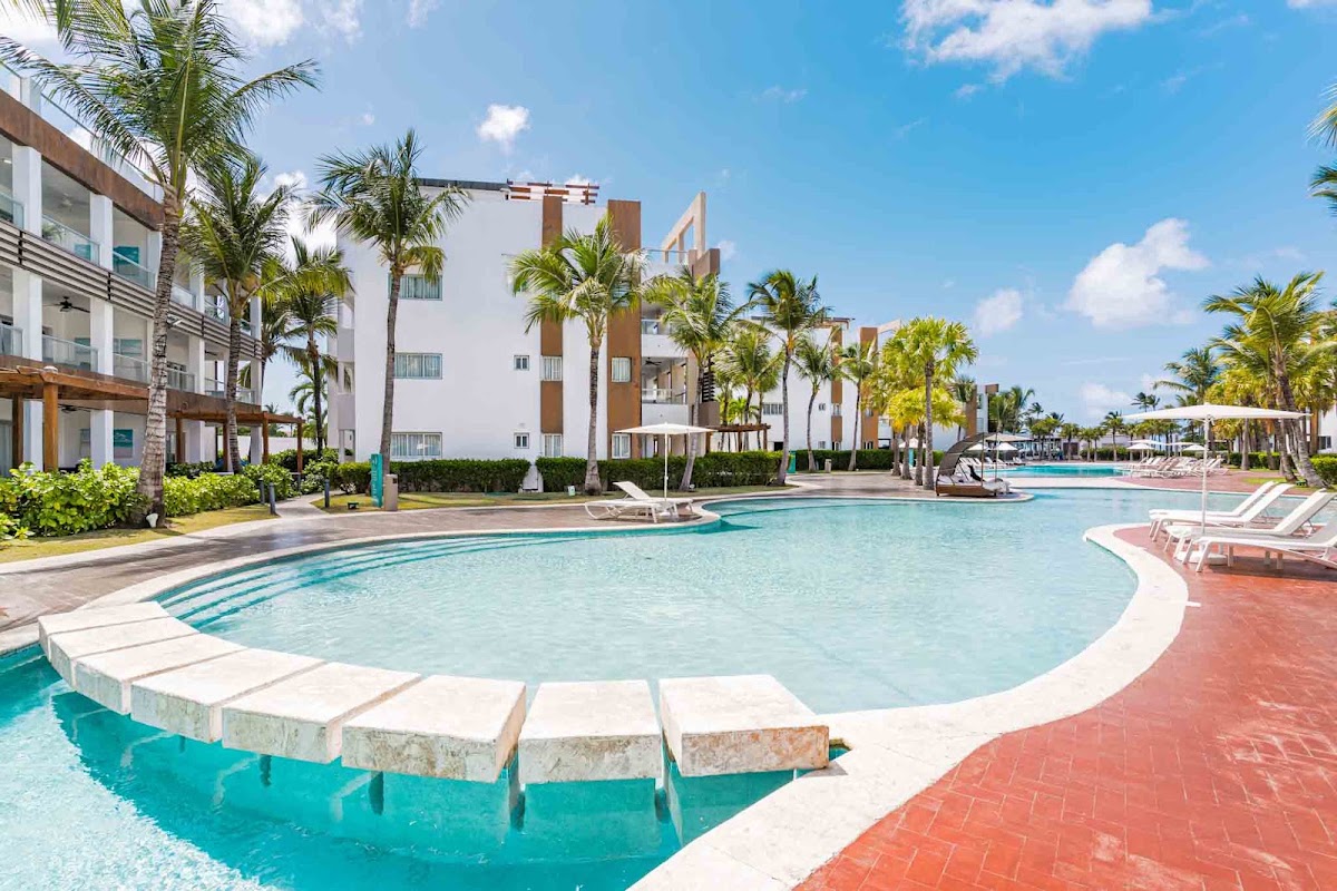 Modern white resort buildings with brown accents and palm trees surround a curved turquoise pool with loungers and a shallow seating area.