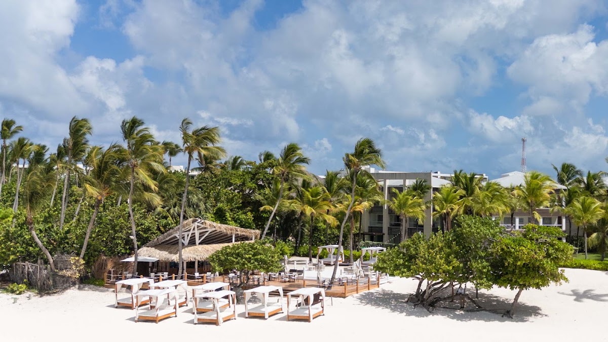 White lounge chairs arranged on white sand beach with thatched-roof cabana and palm trees at Bakour Punta Cana Suites.