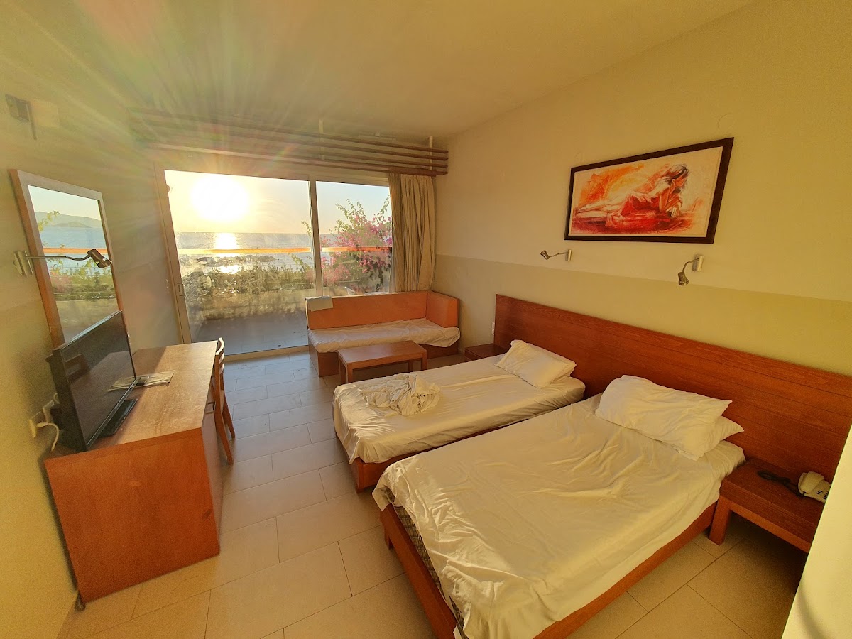 Hotel room with two beds, orange headboards, desk with TV, seating area, and balcony overlooking water at sunset.