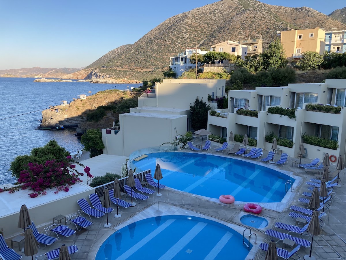 Two blue pools with striped loungers and umbrellas overlook a rocky coastline and blue sea at a cliffside resort in Greece.
