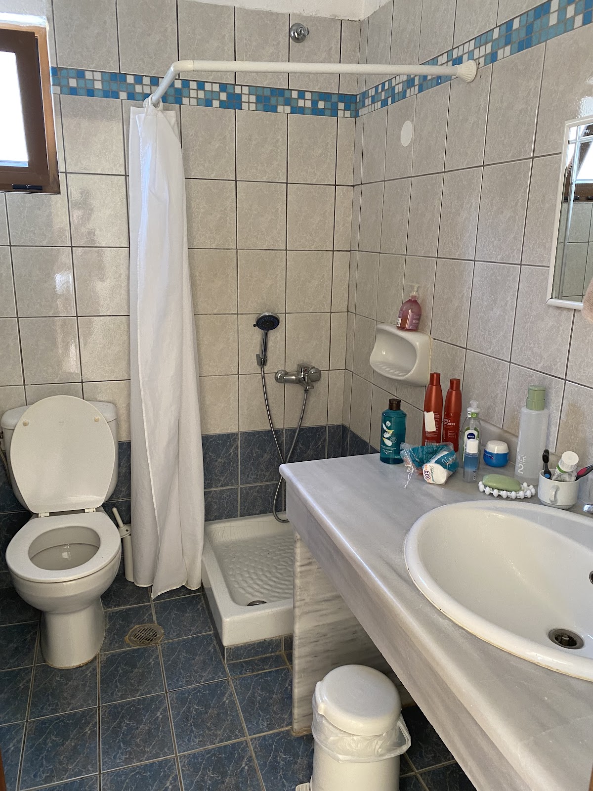 Bathroom with beige tile walls, blue mosaic border, toilet, sink, shower stall with white curtain, and toiletries on counter.