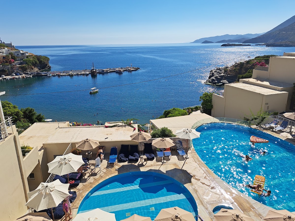 Resort pools and terraces overlook a bright blue bay with boats and rocky coastline under clear sky.