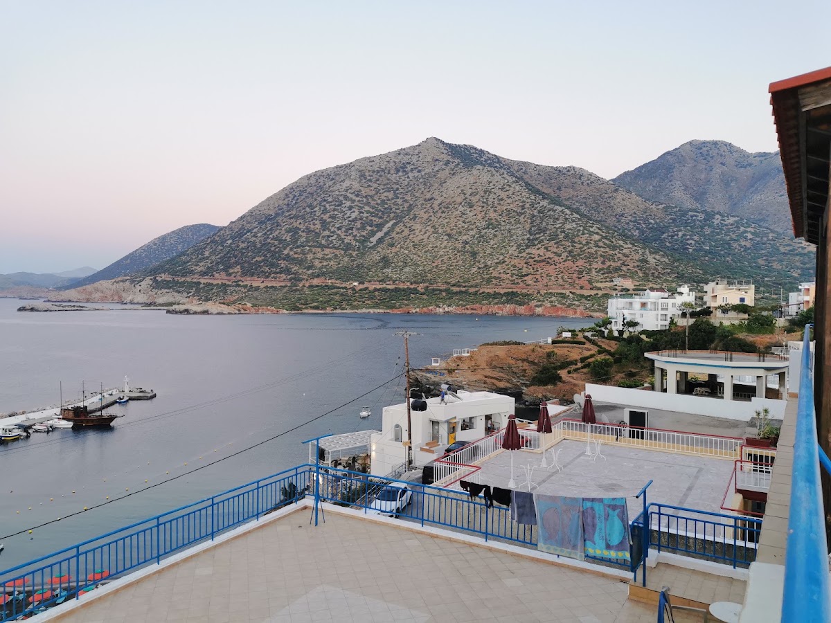 Terraced resort decks with blue railings overlook a bay with fishing boats, rocky coastline, and arid mountains beyond.