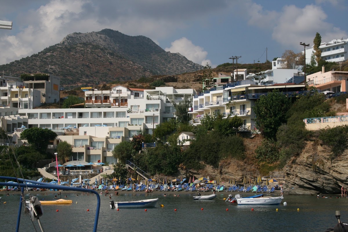 Coastal village with white and pastel buildings built into a hillside above a beach lined with blue umbrellas and boats on the water, rocky mountain in background.