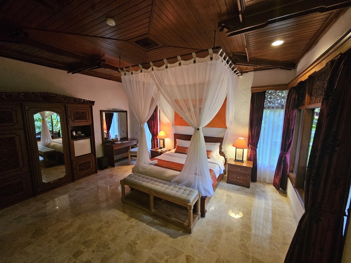 Spacious resort bedroom with a four-poster canopy bed draped in white curtains, wooden ceiling, nightstands with lamps, and access to adjoining rooms.