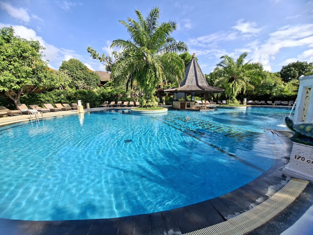 Large blue pool with central thatched-roof bar pavilion, lounge chairs along edges, palm trees and lush gardens surrounding the Bali Tropic Resort & Spa.