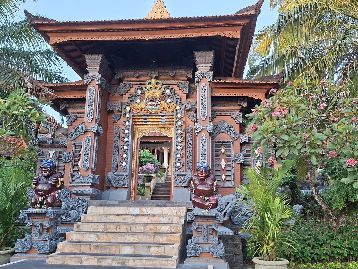 Ornate Balinese temple structure with carved wooden details, stone guardian statues, and tropical flowering plants flanking the entrance.