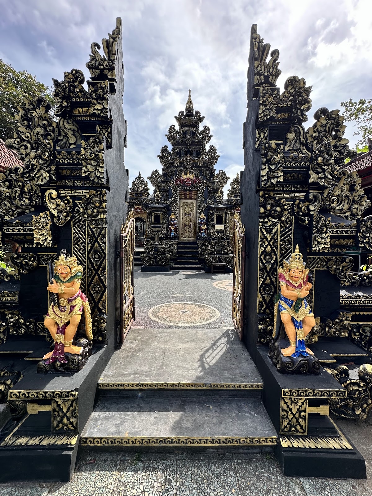 Ornate black and gold temple gates with carved guardian statues frame a view of an intricately decorated Balinese temple at Bali Tropic Resort & Spa.