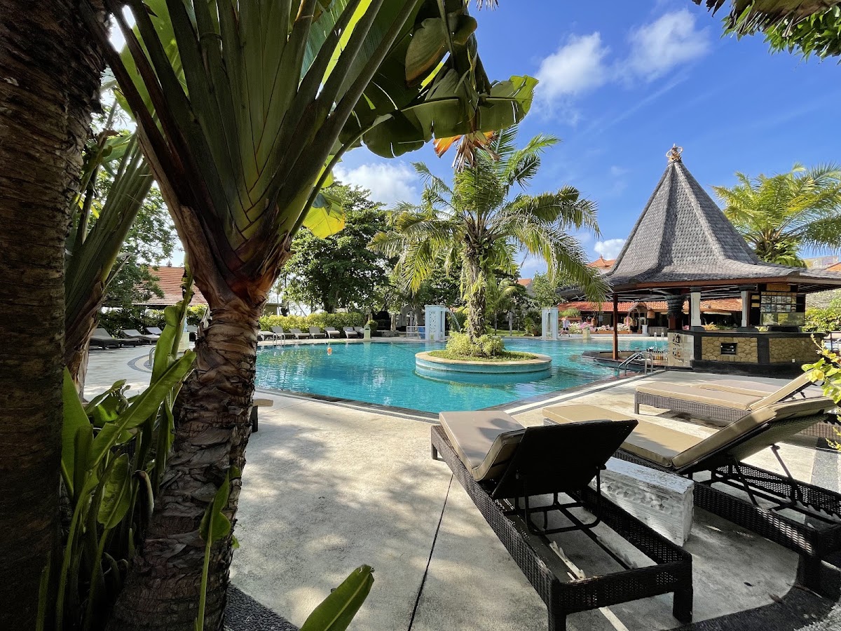 Large turquoise pool surrounded by palm trees and lounge chairs at Bali Tropic Resort & Spa, with a peaked-roof bar pavilion in the background.