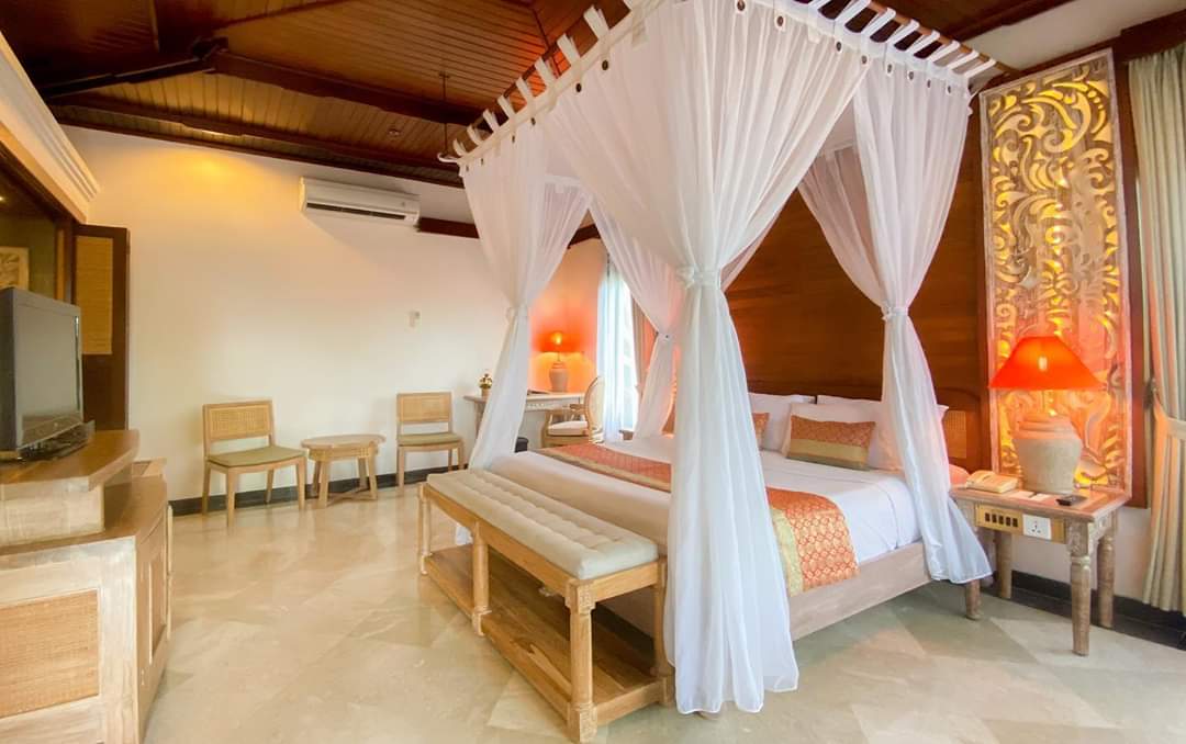 Spacious bedroom at Bali Tropic Resort & Spa with four-poster canopy bed draped in white curtains, wooden furnishings, and warm ambient lighting.