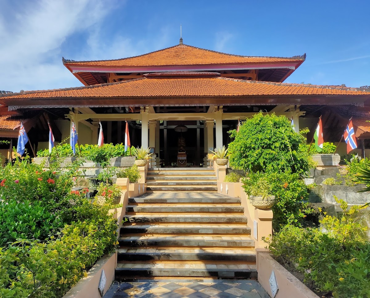 Asian-style pavilion with tiered orange tile roofs, cream-colored columns, and flags flanking a central staircase lined with green plants and flowers.