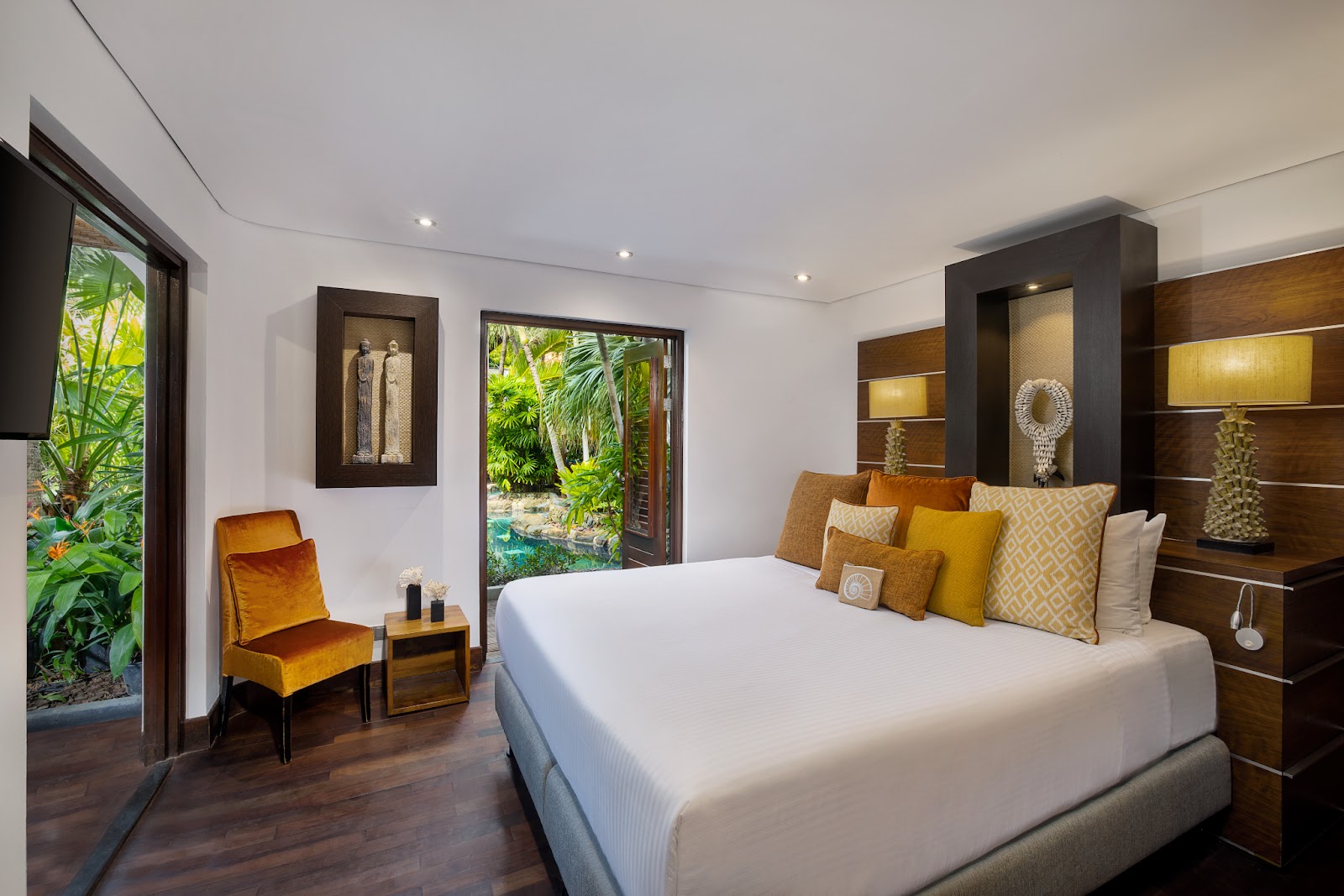 Modern bedroom at Baoase Luxury Resort with white bed, gold velvet chair, dark wood built-ins, and tropical garden views through glass doors.
