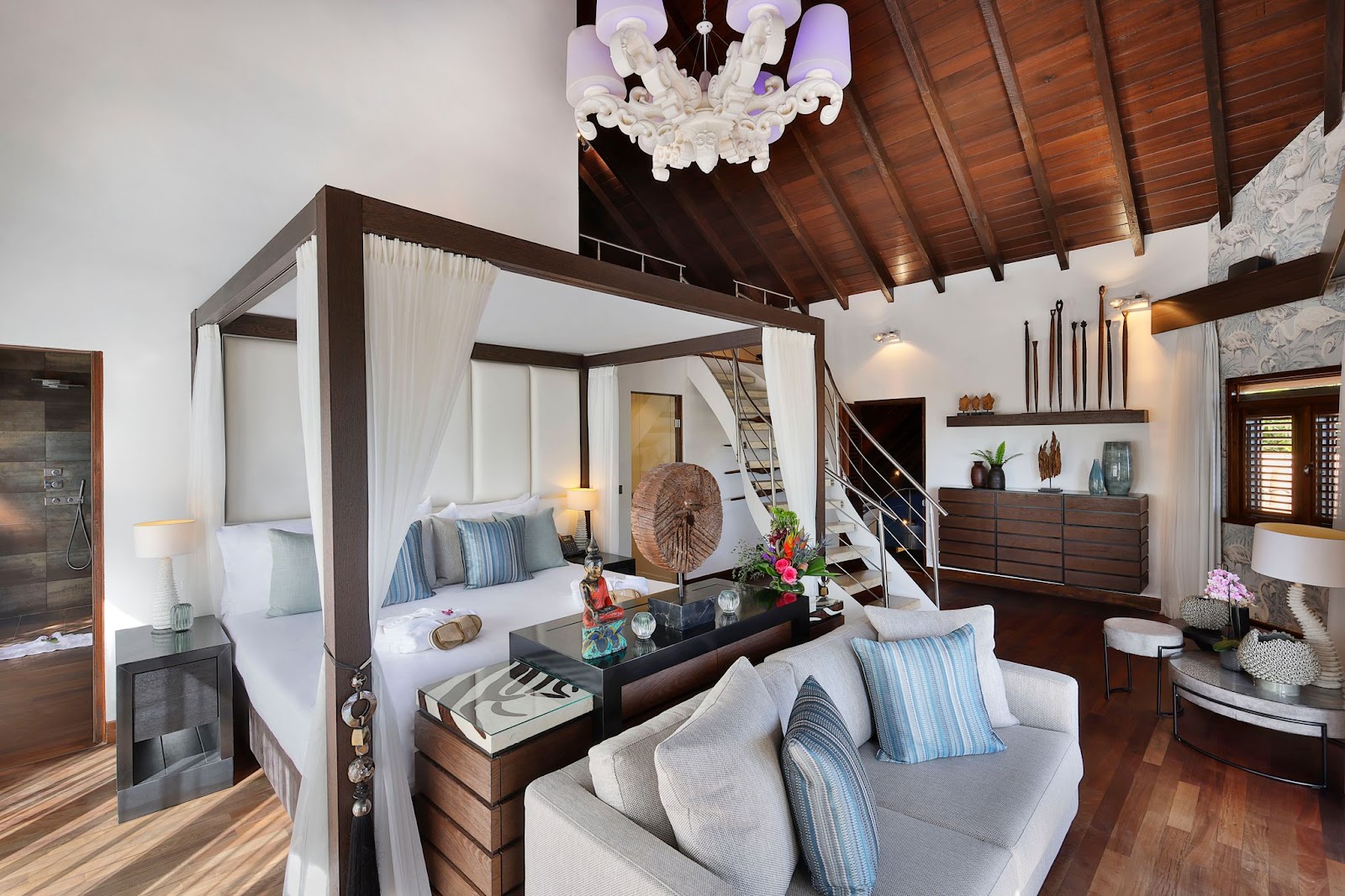 Spacious resort suite with four-poster bed, white curtains, wooden beams, cream sofa, dark wood furnishings, and decorative chandelier.