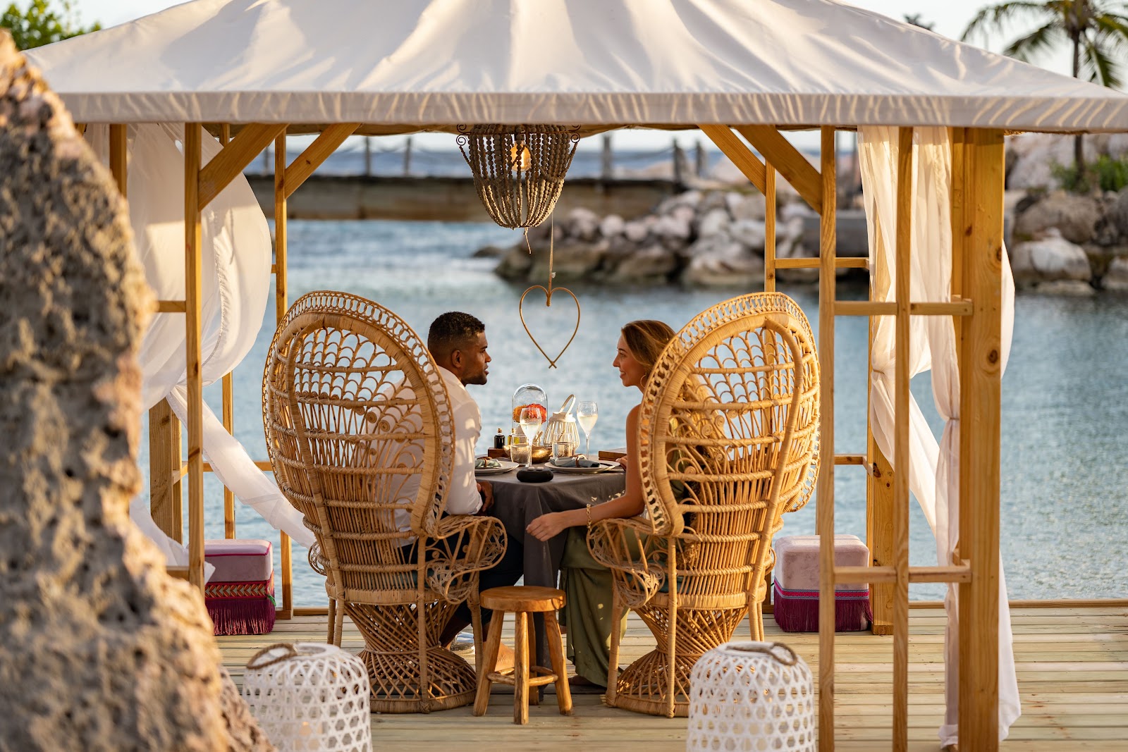 Couple dining at a table under a wooden pavilion with cream canvas roof and hanging heart decoration, overlooking the ocean at Baoase Luxury Resort.